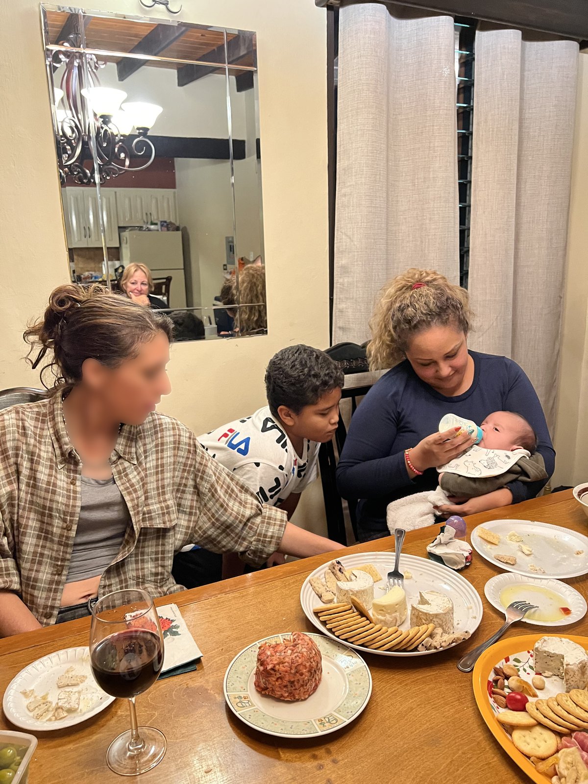 A woman in a brown plaid shirt sits at a candlelit dinner table set with a cheese board and crackers, a young person leaning beside her, while another woman in dark blue holds a baby feeding from a bottle and a third adult is visible in the wall mirror behind them.