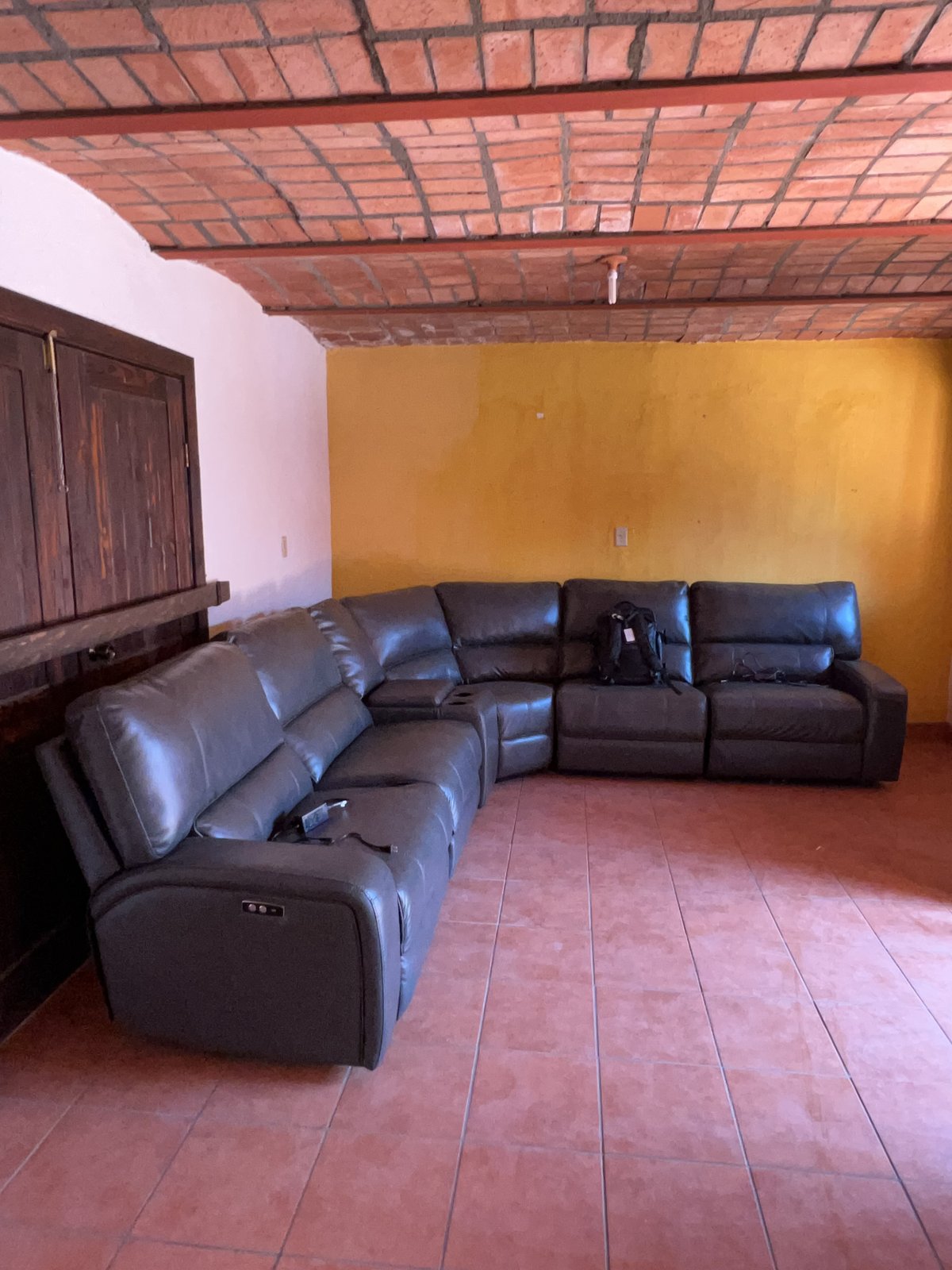 An empty room shows a curved brick bóveda ceiling overhead, a long grey leather sectional sofa along bright yellow walls, dark wood cabinetry on the left, and terracotta tile flooring.