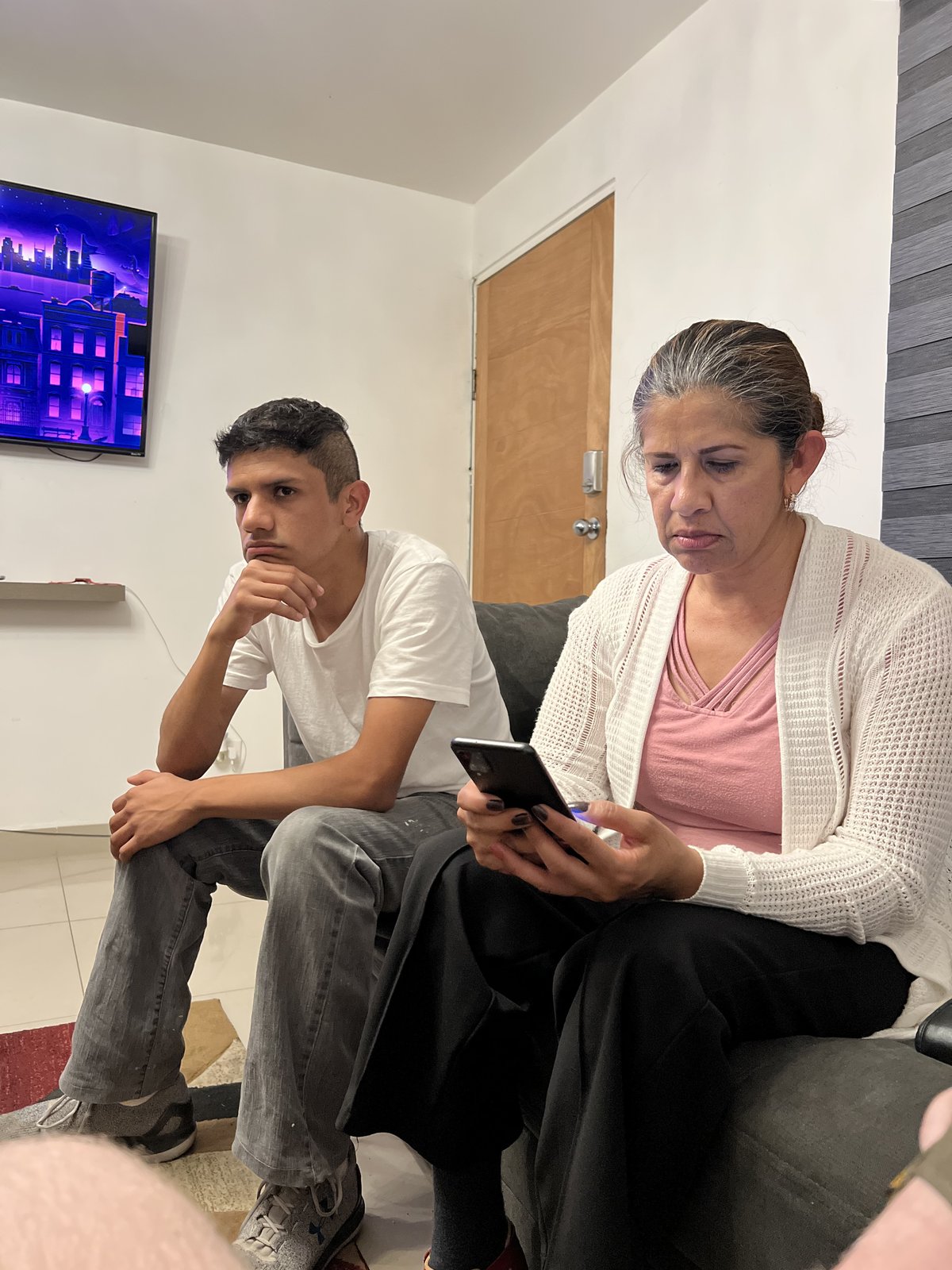 A young man in a white t-shirt sits on a couch with his hand at his chin beside an older woman in a cream knit cardigan and pink top who looks down at a phone in her hand, a wall-mounted television showing a purple cityscape behind them.