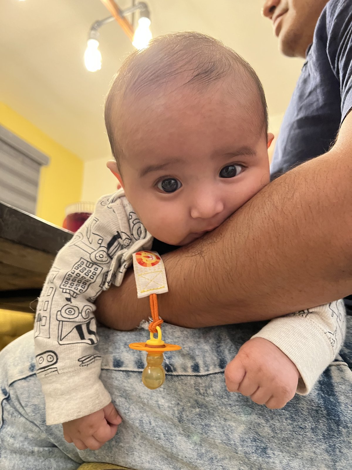 A wide-eyed baby in a printed long-sleeved onesie leans against an adult's bare forearm, an orange pacifier on a fabric tether resting on the baby's wrist, with overhead lightbulbs and a yellow wall behind.