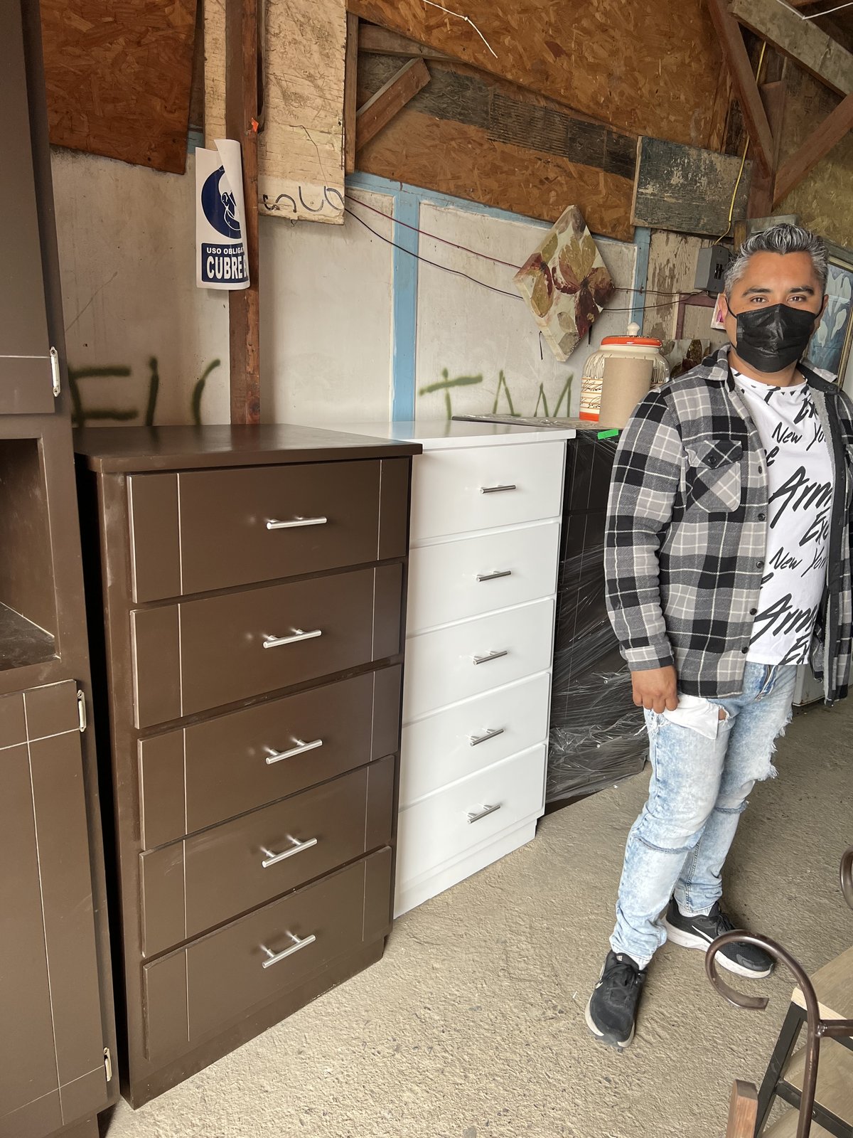 A man in a checkered flannel shirt and a black face mask stands beside a brown and a white five-drawer dresser inside an open wood-frame warehouse with graffiti on the back wall.