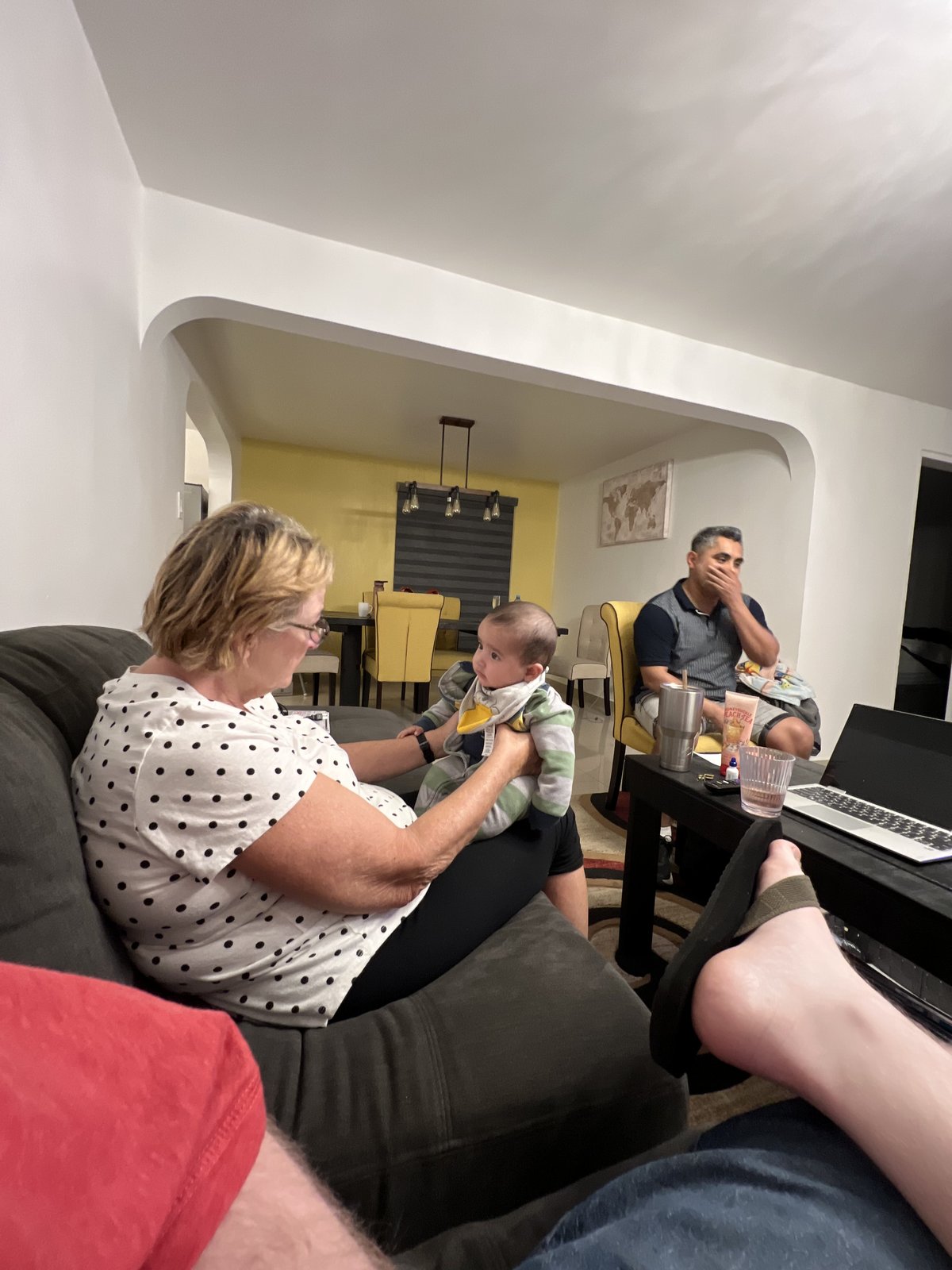 A woman in a black-and-white polka-dot top sits on a dark grey sofa cradling a baby in striped clothing, while across a living room a man in a dark polo sits beside a laptop and a glass of water, with another person's legs visible in the foreground.