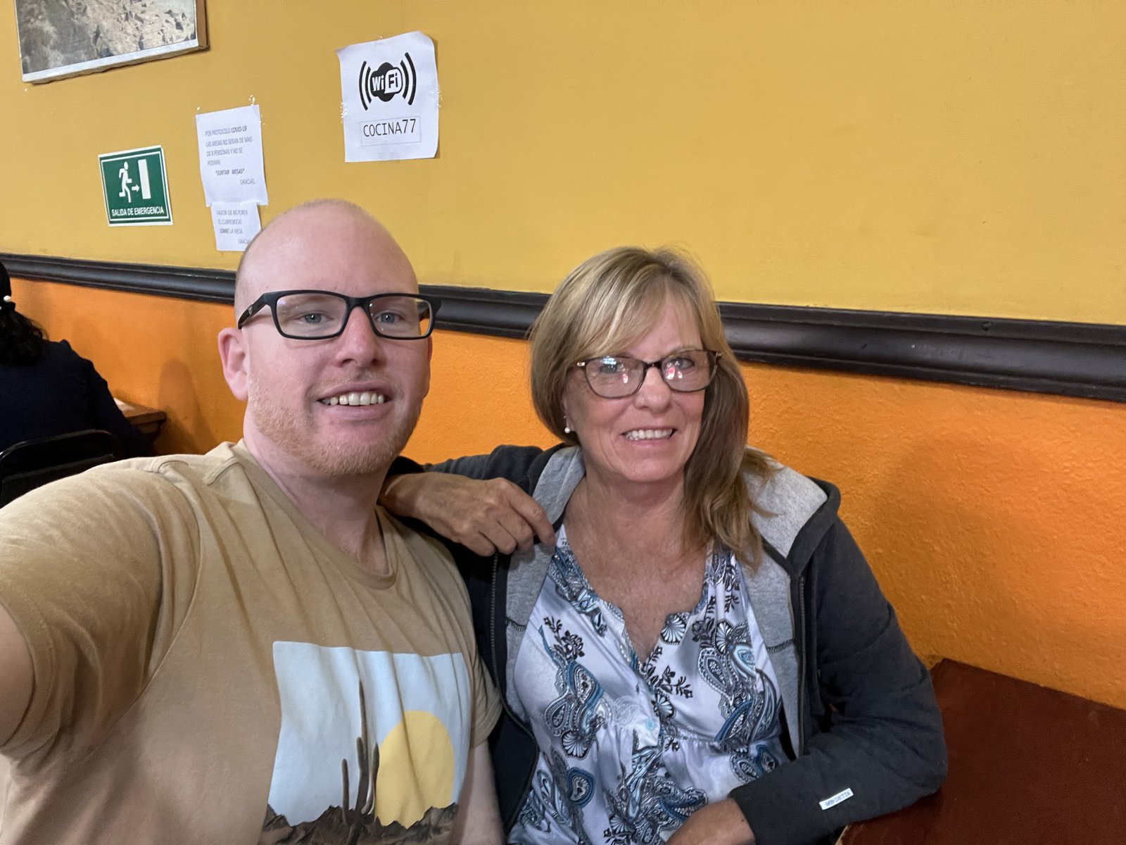 A bald man in glasses and a t-shirt and a smiling blonde woman in glasses pose closely together for a selfie in front of a yellow wall displaying a printed 'wifi COCINA77' sign and a green emergency exit sign.